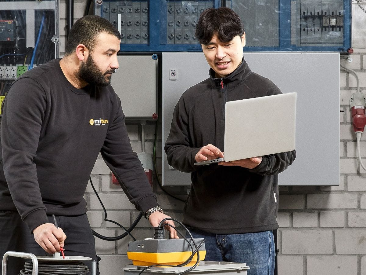 Zwei Mitarbeiter bei der Geräteprüfung: einer misst am Prüfgerät, der andere trägt das Ergebnis direkt am Laptop ein.