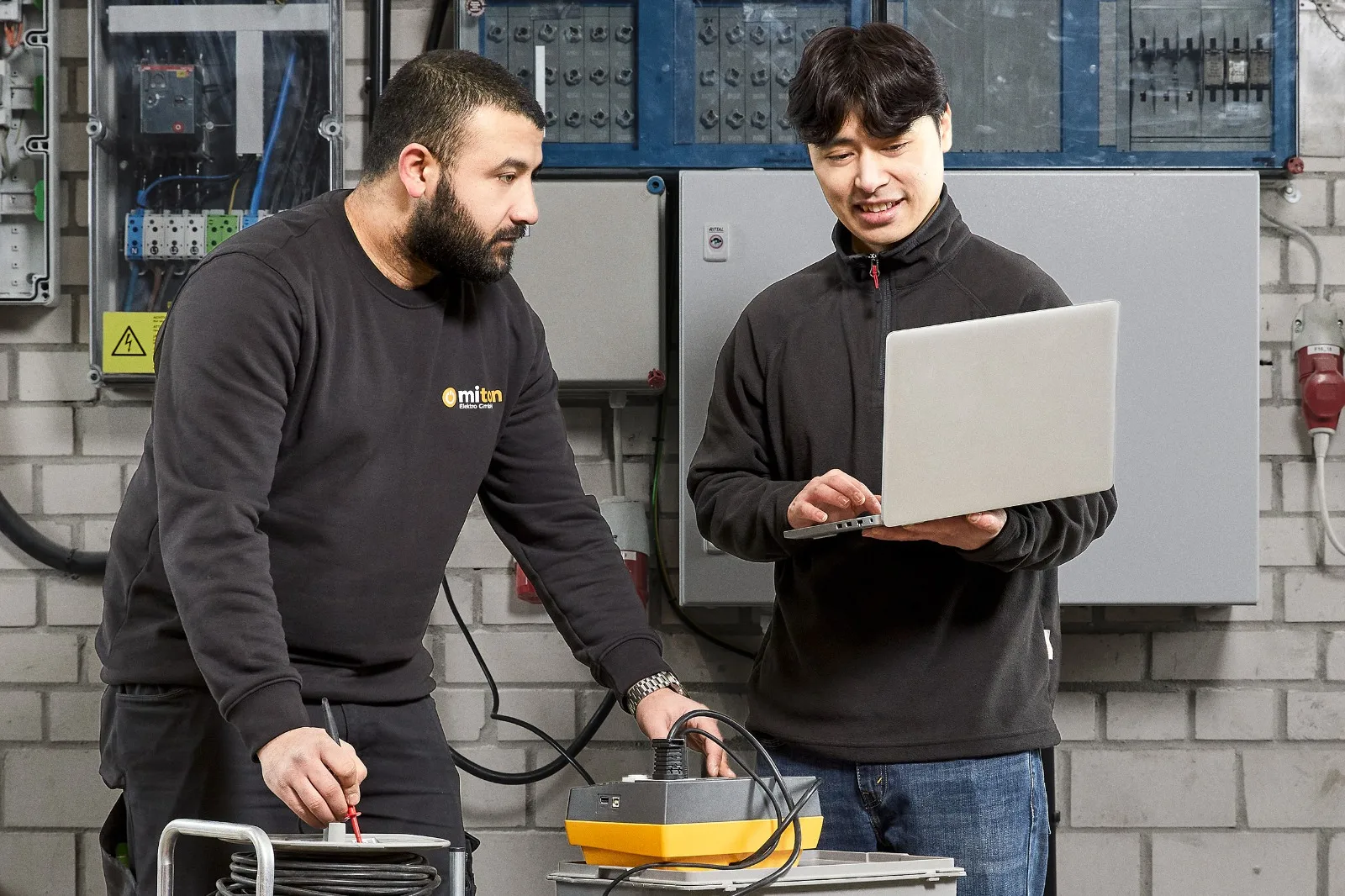 Zwei Mitarbeiter von Miton Elektro bei der Geräteprüfung: einer misst am Prüfgerät, der andere trägt das Ergebnis am Laptop ein.